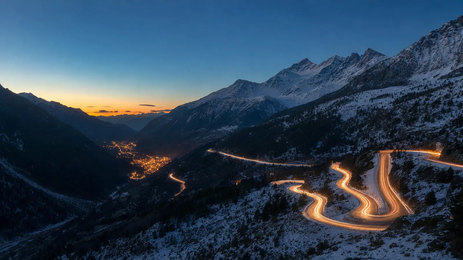 Route de col enneigée des Pyrénées à l’heure bleue, traînées lumineuses de voitures vers la vallée